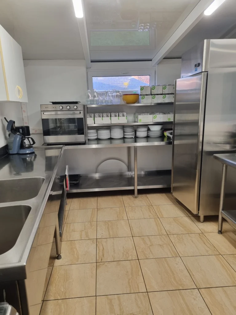 Cuisine de la salle des fêtes : fours, vaisselle sur étagères et réfrigérateur.