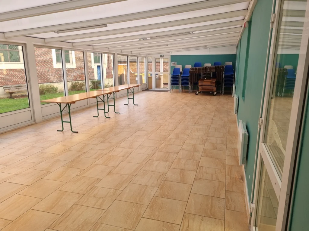 Véranda vitrée de la salle des fêtes avec des tables et des chaises empilées.