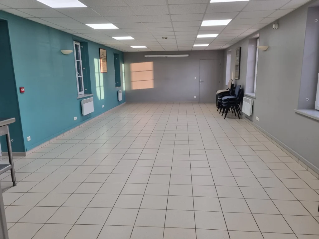 Véranda vitrée de la salle des fêtes avec des tables, ouverte sur la cour.
